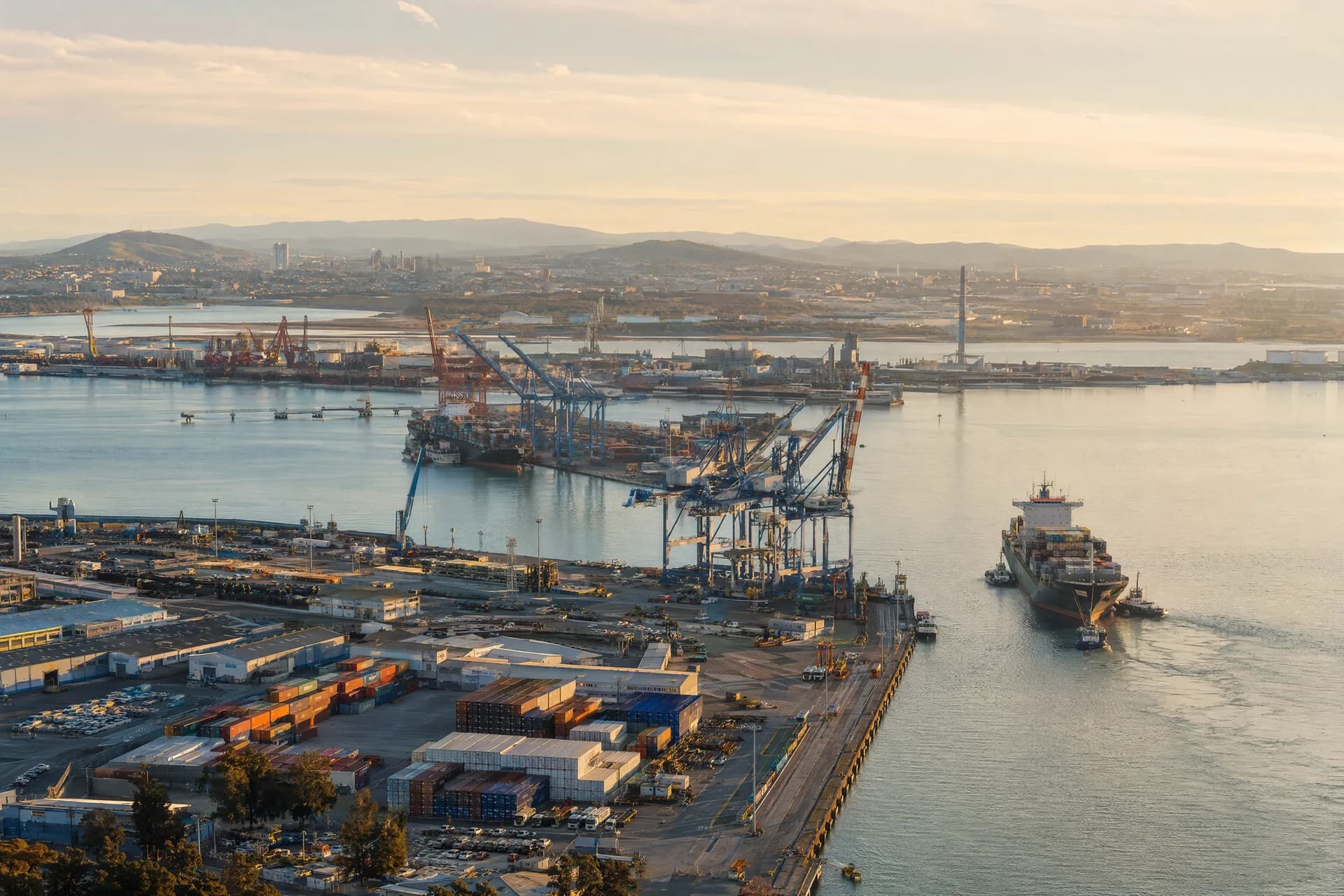 Elevated view of a container port with cranes, warehouses and a cargo ship assisted by tugboats.
