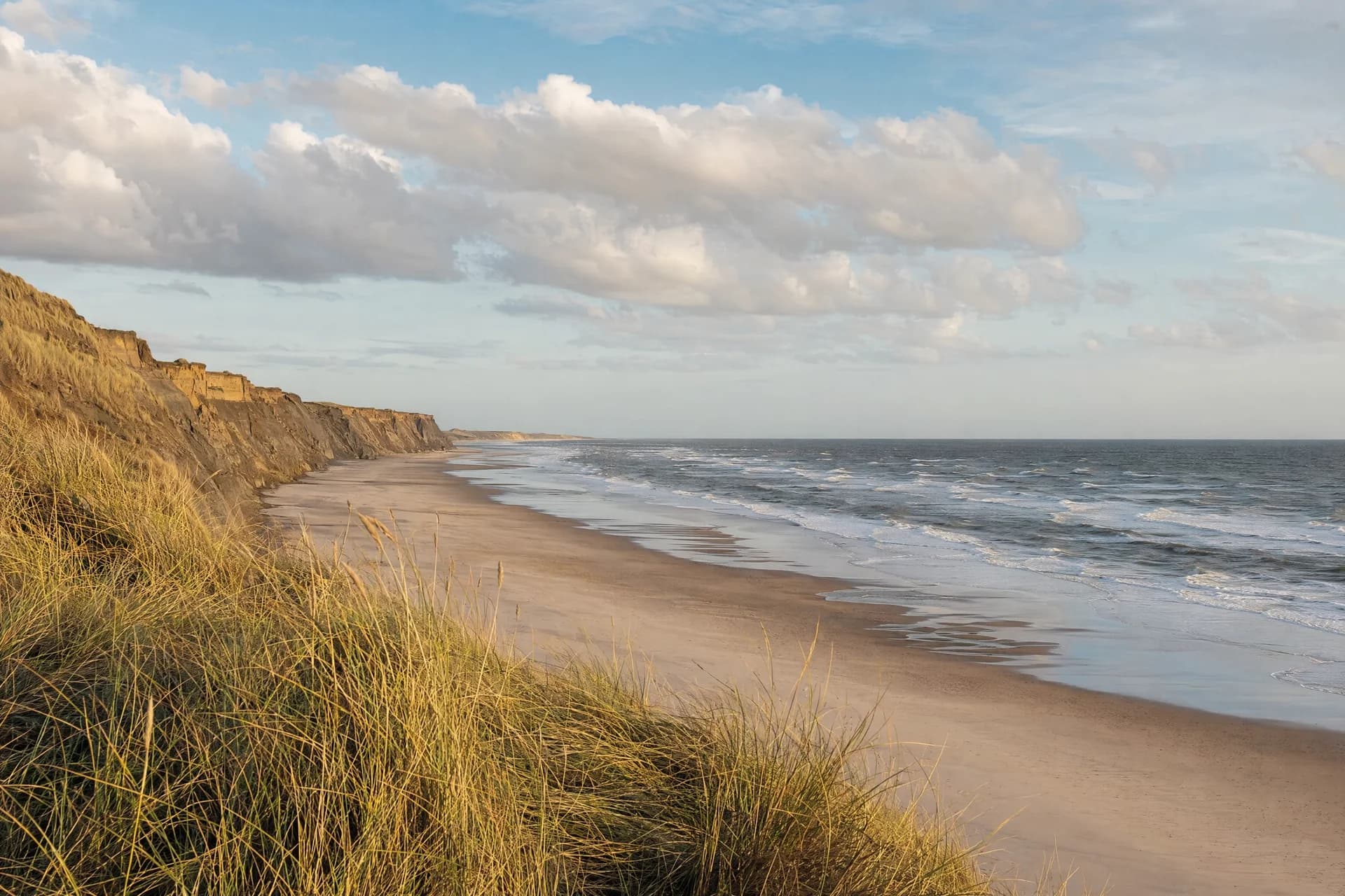 Sandy coastline beneath grassy cliffs, with waves breaking along the shore in warm light.