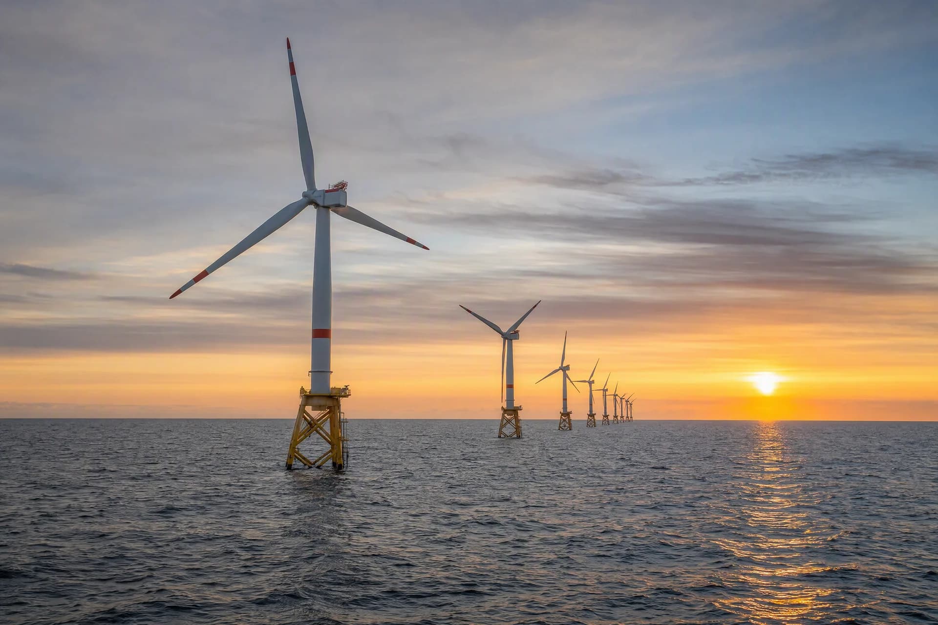 Offshore wind turbines on yellow lattice foundations receding across the sea at sunset.