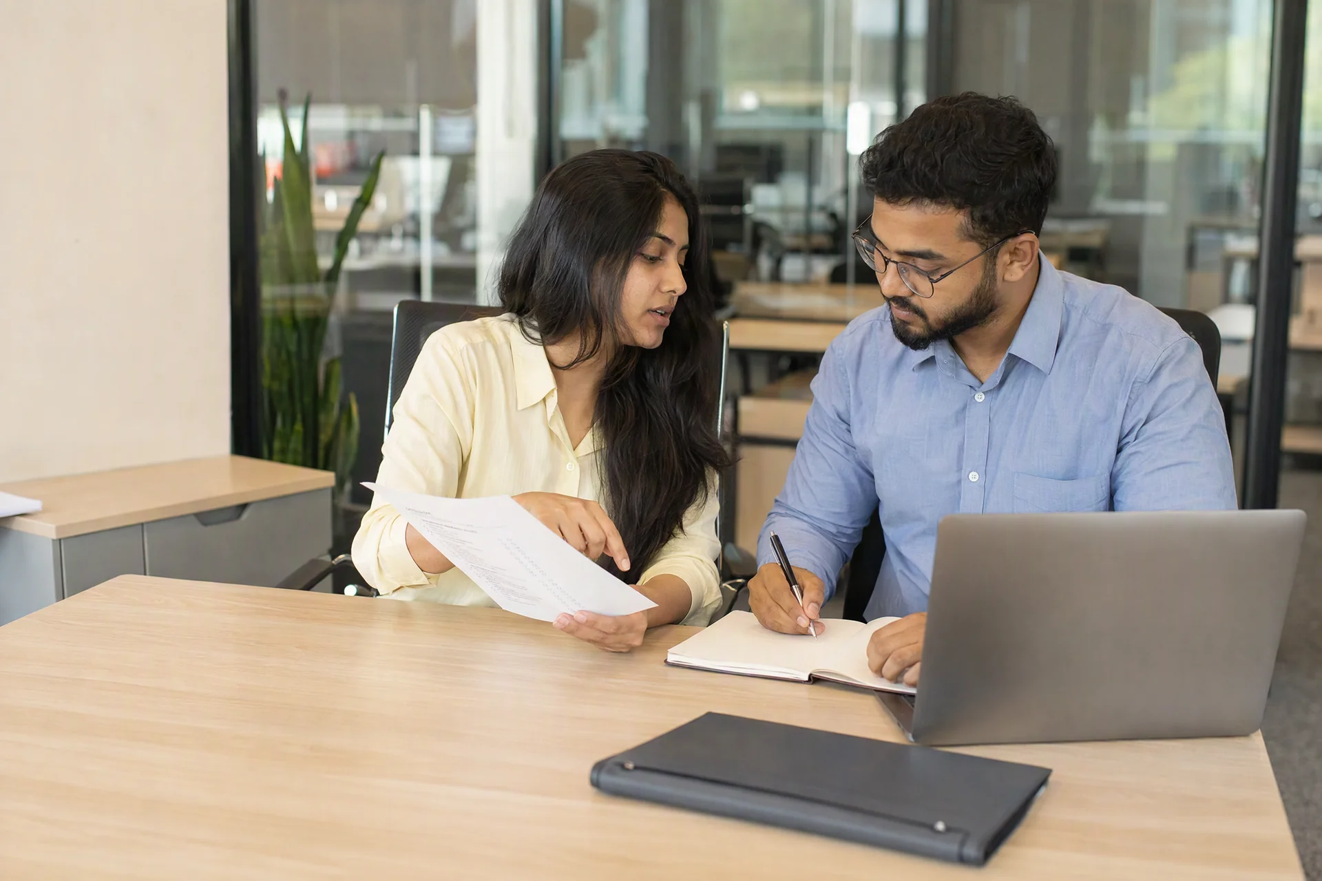 Two colleagues reviewing a handover checklist and taking notes beside a laptop in a working office.