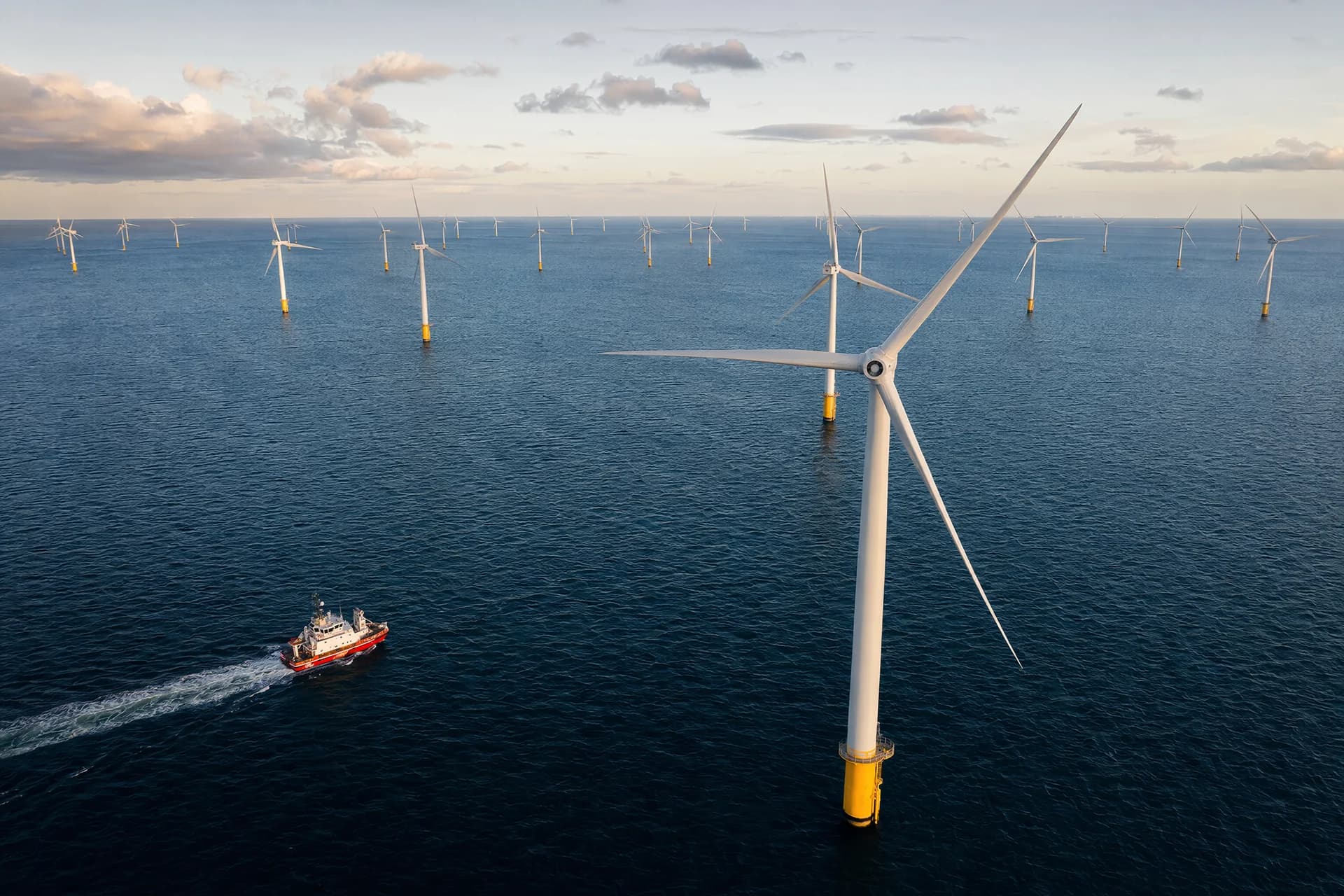 Aerial view of offshore wind turbines on yellow foundations, with a small workboat crossing the sea below.
