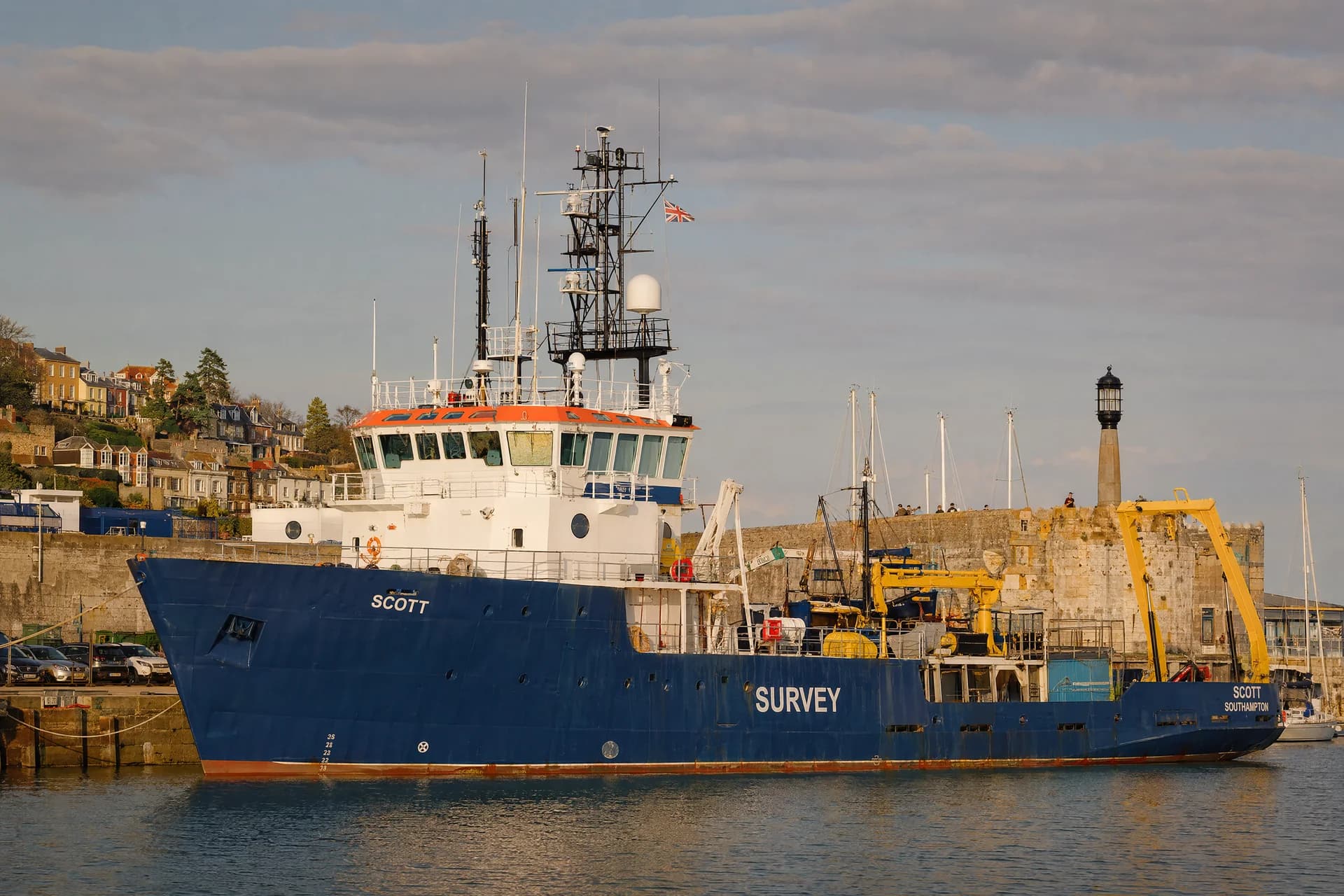 Blue and white survey vessel moored beside a stone harbour wall in warm sunlight.